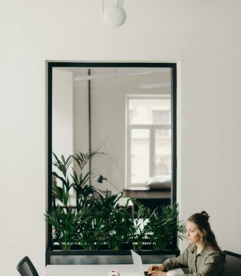 A young woman working on a laptop in a minimalistic, modern office setting with plants and a cup of coffee.